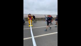 Stormy Stan (RNLI mascot) holds a ribbon as a runner approaches a finish tape holding an RNLI200 flag