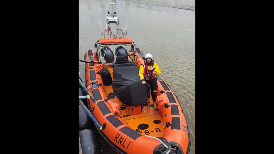 A man in a white helmet, yellow drysuit, and red lifejacket stands on an orange Atlantic 85 lifeboat.
