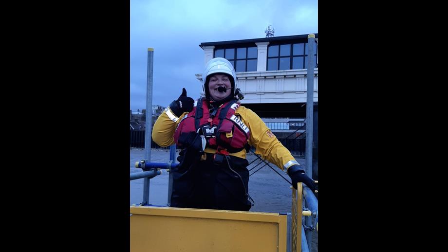 A woman in a white helmet, yellow drysuit, and red lifejacket smiles and gives a thumbs up to the camera
