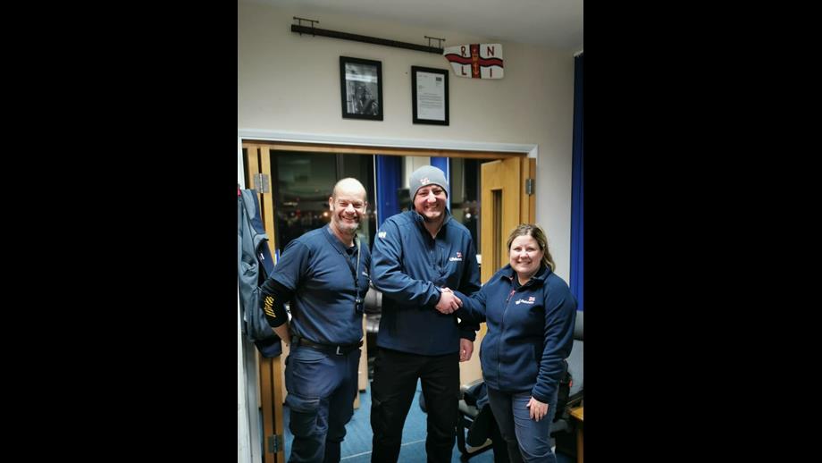 Three people in navy RNLI branded clothing stand indoors smiling at the camera. The man in the centre shakes the hand of the woman on the right.
