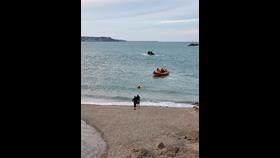 A crew member makes their way ashore from the lifeboat towards two members of the public standing on teh shore. The crew member is chest deep in the water as the lifeboat stands off.