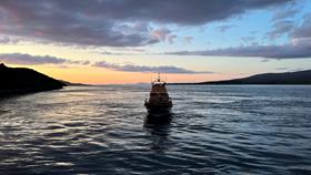 Islay RNLI lifeboat sits in the water, centre of the image, bow facing the camera. There is a dusky, yello and pink sunrise behind.