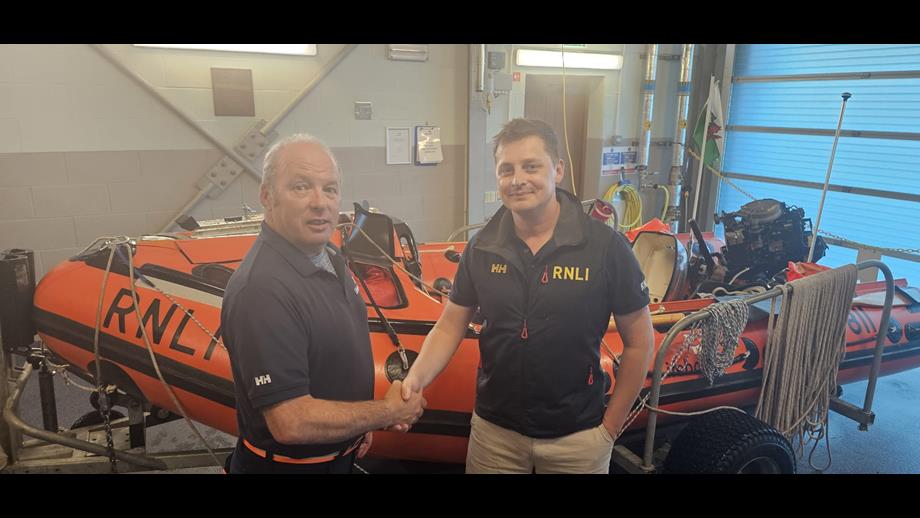 Anton Hook (right) Pwllheli RNLI D class inshore lifeboat helm shaking hands with RNLI trainer/assessor Simon Bunting (left). They are standing in front of a D-Class inshore lifeboat at Pwllheli lifeboat station.
