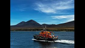 Islay lifeboat, a large all-weather lifeboat, underway to a yacht. There is white water behind the boat and the background it the paps of Jura, a server of iconic large hills on the opposite island. The weather is sunny with a blue sky.