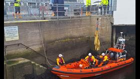 Inshore lifeboat in the water alongside the quay wall after returning to the lifeboat station. Two crew are on board and a third is standing alongside on the steps to the station. Just out of sight are two shore crew holding lines to the lifeboat.