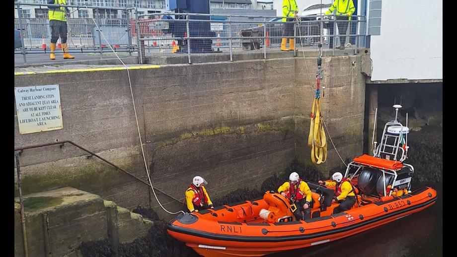 Inshore lifeboat in the water alongside the quay wall after returning to the lifeboat station. Two crew are on board and a third is standing alongside on the steps to the station. Just out of sight are two shore crew holding lines to the lifeboat.
