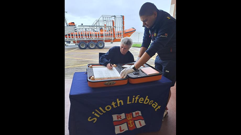 Female sitting on chair behind table signing scroll with orange lifeboat in the background