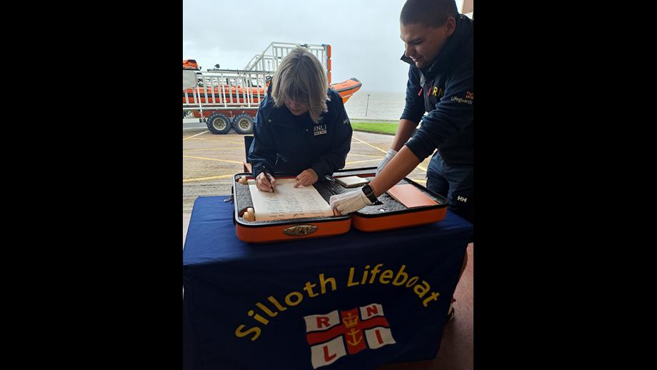 Female standing behind table signing scroll with orange lifeboat in the background
