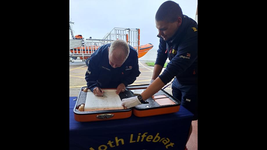 Male sitting on chair behind table signing scroll with orange lifeboat in the background