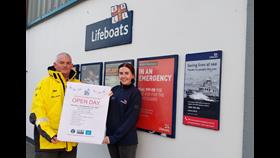 Two crew holding a poster for the open day, just visible in the background are the poster display cases on the outside of the lifeboat station