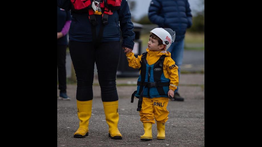 Child dressed in yellow clothing and yellow wellie wearing blue life vest holding mother's hand looking to lifeboat beside them
