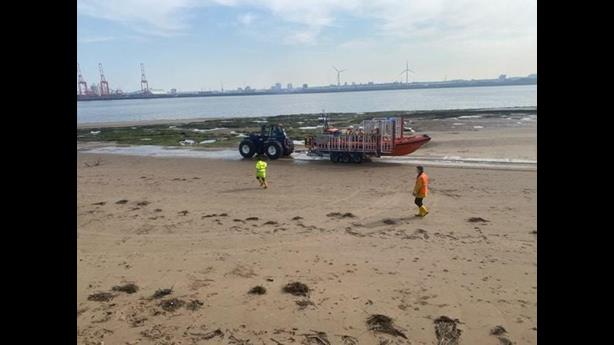 New Brighton beach, with shore crew visible in hi-vis gear escorting the Bendy tractor, carriage and lifeboat to the water's edge. In the background the Mersey can be seen.