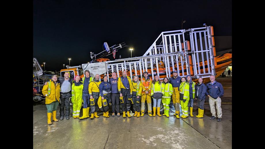 Night time, a large group of New Brighton RNLI volunteers pose in front of their lifeboat, held on the carriage, outside the boathouse.
