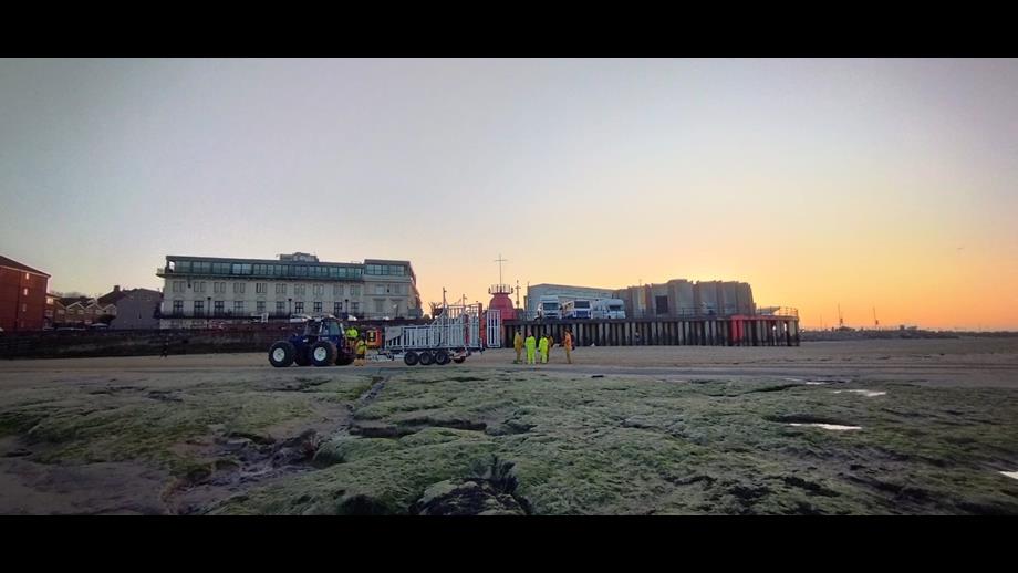 A wide angle shot from a distance. Sunset, with beautiful golden colours in the sky. Silhouetted against the beach and the old buildings beyond, New Brighton RNLI's tractor, carriage and lifeboat can be seen moving up the beach, with shore crew either side, escorting.