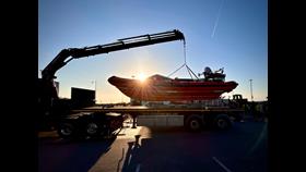 A beautiful sunset sky in New Brighton. Silhouetted against the sunset the lifeboat can be seen, attached to a crane arm, being lifted from the back of a flat bed truck.
