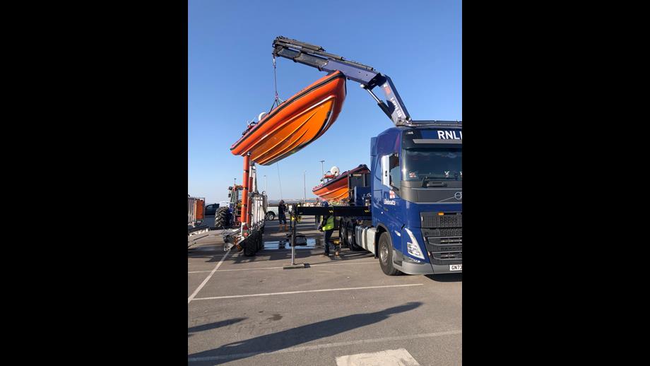 The B-class lifeboat can be seen suspended in mid-air, lifted by crane arm, as it is moved out of the now empty carriage to the left of the frame, to the flat bed truck on the right.