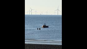 A portrait shot of the Mersey, with wind farms visible in the background, and beach in the foreground. Two Coastguard rescue units head out, wading in the water, to greet New Brighton RNLI's Atlantic-85, with the casualty vessel.
