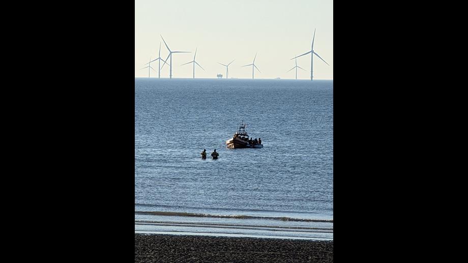 A portrait shot of the Mersey, with wind farms visible in the background, and beach in the foreground. Two Coastguard rescue units head out, wading in the water, to greet New Brighton RNLI's Atlantic-85, with the casualty vessel.
