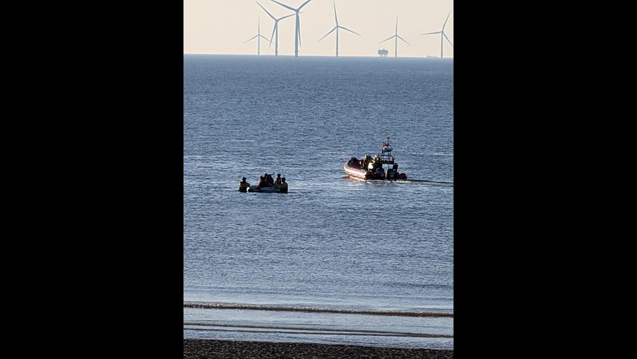 Wind farm in the background, small strip of beach in the foreground. The majority of the frame is dominated by water, in a portrait orientation, as New Brighton RNLI head away from the scene. Two Coastguard units can be seen assisting the small dinghy of casualties back to shore. 