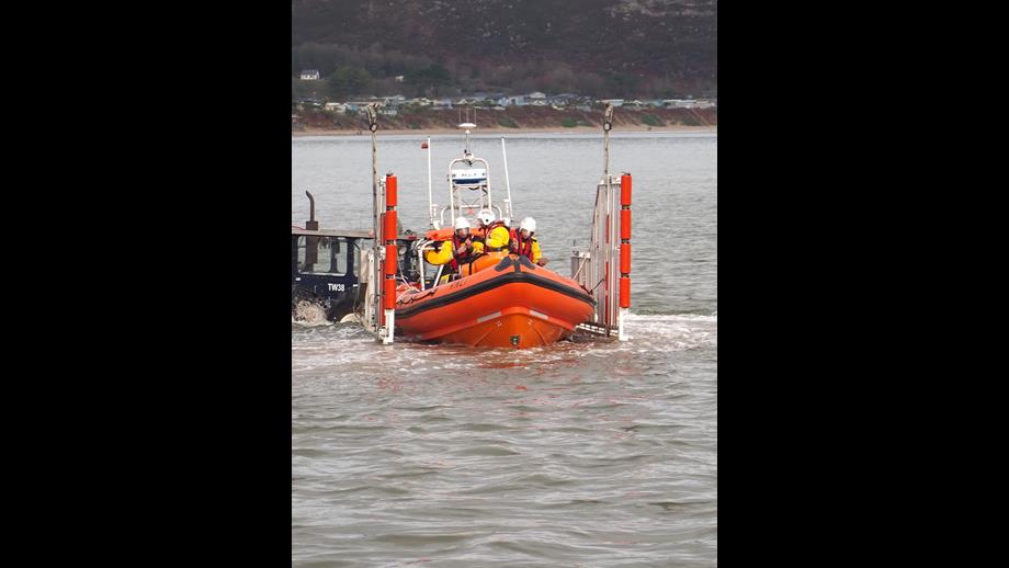 A lifeboat carrying several crew members in yellow and red uniforms is being deployed into the water from a vehicle on a ramp. The scene takes place near the shore with hills and buildings visible in the background.
