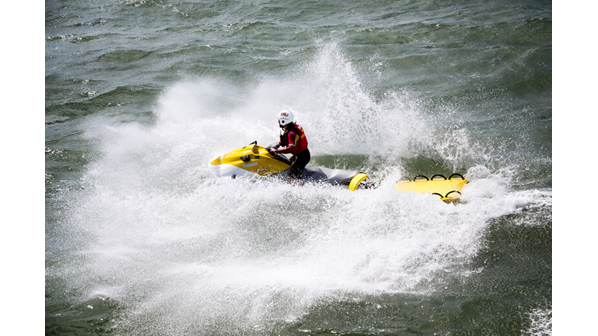 Lifeguard wearing a white helmet on a yellow and white rescue water craft crashes through the waves with a yellow rescue board attached.