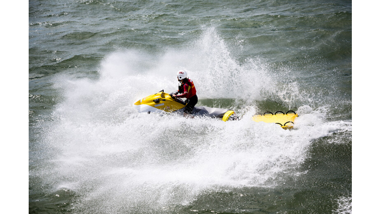 Lifeguard wearing a white helmet on a yellow and white rescue water craft crashes through the waves with a yellow rescue board attached.