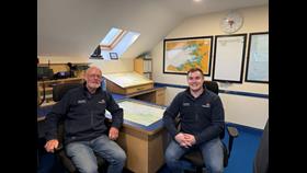Two crew wearing navy RNLI jackets sitting in the ops room of Galway Lifeboat Station with charts and comms equipment in the background