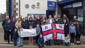 Standing outside Kilmore Quay RNLI station, a group of adults made up of RNLI volunteers and fundraisers along with children in the front some holding a large size cheque and others a large RNLI flag