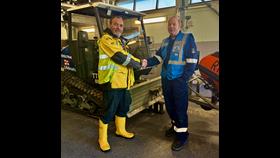 Dave Hill on the left wearing RNLI high vis jacket, shaking hands with Simon Bunting, RNLI assessor. They are standing in front of a ToolTrack tracked launch vehicle.