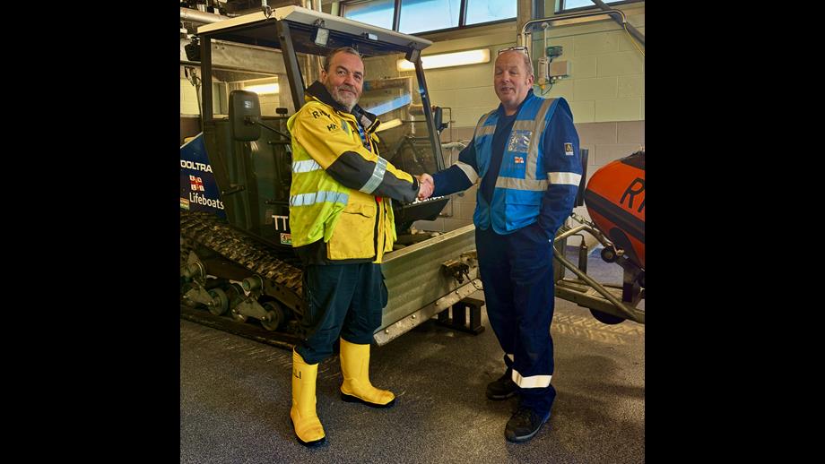 Dave Hill on the left wearing RNLI high vis jacket, shaking hands with Simon Bunting, RNLI assessor. They are standing in front of a ToolTrack tracked launch vehicle.