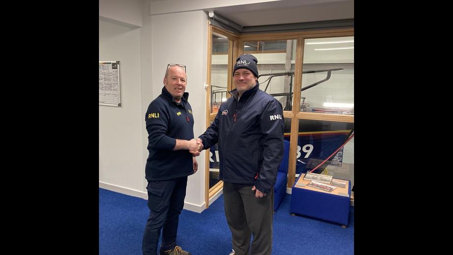 Simon Buntin on the left, shaking hands with Paul Kitteingham on the right. The all-weather lifeboat can be seen in the background through the windows.