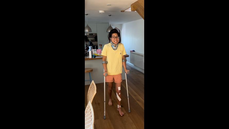 Vicki pictured at home using crutches and smiling wearing a neck brace and with dressings on her left leg.