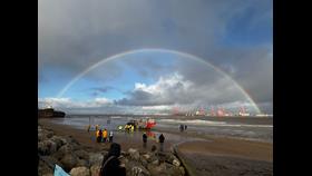 Landscape image of New Brighton beach. Volunteer crew in yellow can be seen on the sand, with the tractor, carriage and lifeboat just beyond them. A rainbow stretches across the sky, from the Fort on the left of the frame, to the Liverpool docklands on the right of frame. 