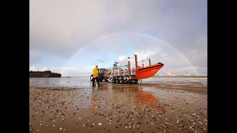 Low shot of New Brighton beach, with the Fort Perch Rock visible to the left of the frame. In the centre is the lifeboat on the carriage, with a single volunteer walking alongside. A rainbow stretches across the top of the carriage, tractor and lifeboat, framing it. 