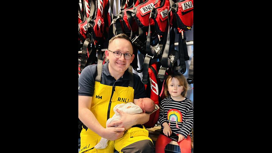 The photo shows a family of 3 - dad in RNLI yellows with his young daughter and baby son in front of some lifejackets