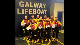 Nine new trainee crew in drysuits and lifejackets standing outside the Galway Lifeboat Station in two rows. The front row are holding helmets.