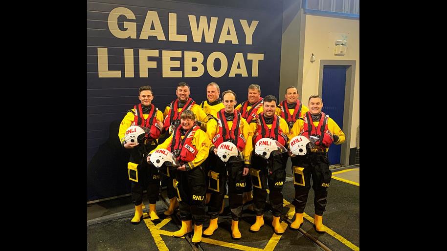 Nine new trainee crew in drysuits and lifejackets standing outside the Galway Lifeboat Station in two rows. The front row are holding helmets.
