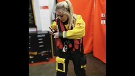 Emily is mid-action, tightening the straps on a red RNLI lifejacket, dressed in her yellow and black dry suit. The background is in soft focus, and is the boathouse at New Brighton.