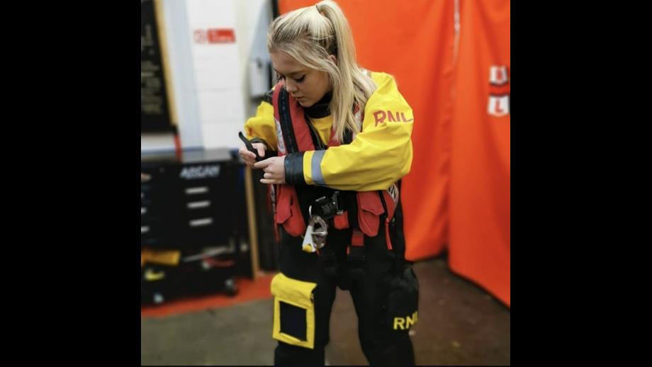 Emily is mid-action, tightening the straps on a red RNLI lifejacket, dressed in her yellow and black dry suit. The background is in soft focus, and is the boathouse at New Brighton.