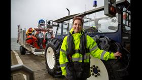 Woman in high visibility jacket stands in front of blue tractor that is towed to orange inshore lifeboat on concrete slipway.