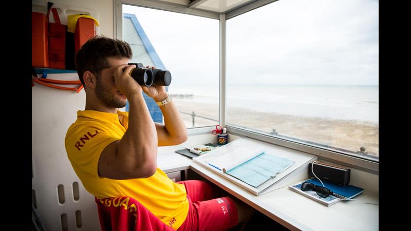 RNLI Lead Lifeguard Supervisor Ted Morgan on watch in yellow and red lifeguard uniform in lifeguard hut on beach