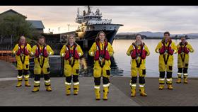 Seven members of the crew in full kit pose in the evening sunshine in front of the Oban lifeboat with the Northern Lighthouse vessel Pharos in the background
