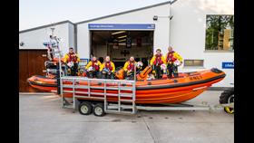 An image showing a group of lifeboat crew members dressed in yellow and red gear sitting on an orange RNLI lifeboat. The boat is mounted on a trailer and positioned outside the Cambyright Lifeboat Station.