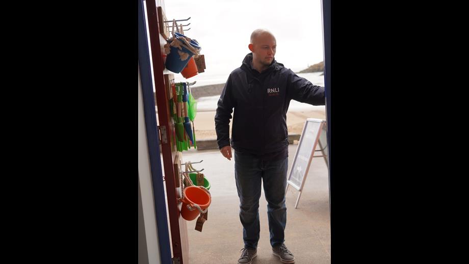 A man stands in the shop doorway, with buckets and spades hanging on the back of the door