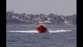 An orange lifeboat moving quickly on the water with waves forming behind it. The background shows a coastal town with various houses and trees.
