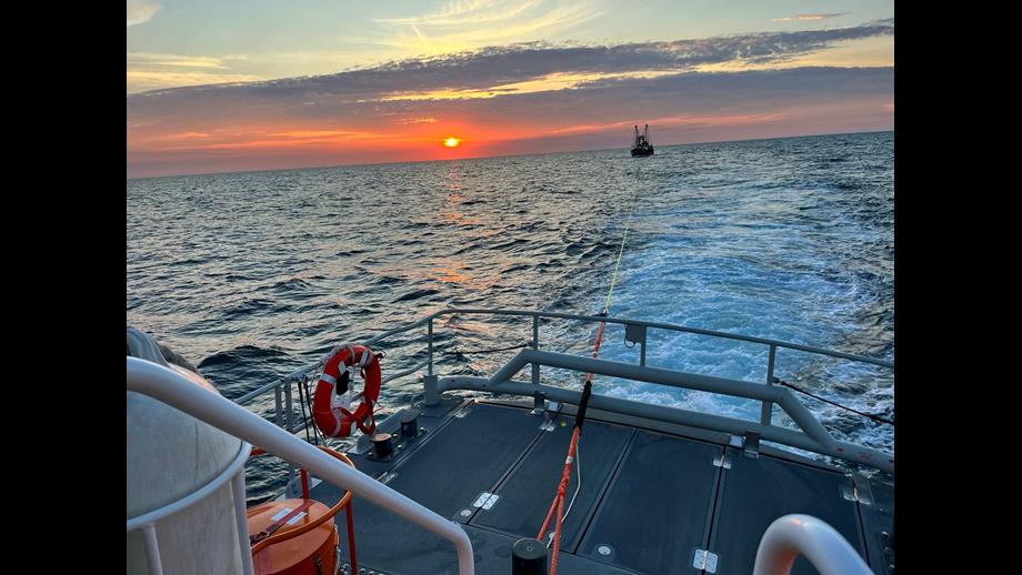 The image shows a beautiful sunset over the ocean with an orange and pink sky. In the foreground, there is a deck of a boat with safety equipment, including a life preserver and ropes. A tow line extends from this boat to another vessel visible in the distance.