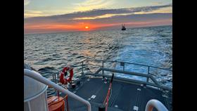 The image shows a beautiful sunset over the ocean with an orange and pink sky. In the foreground, there is a deck of a boat with safety equipment, including a life preserver and ropes. A tow line extends from this boat to another vessel visible in the distance.