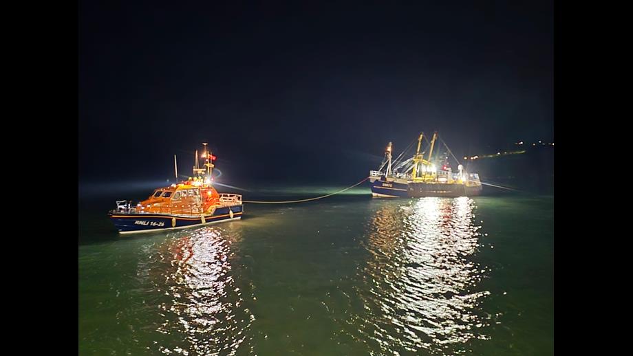 A nighttime scene showing a rescue boat and a fishing vessel on the water. The rescue boat, labeled 'RNLI 14-24,' is brightly lit and connected by a rope to the larger fishing vessel, which is also illuminated. The water reflects the lights from both boats.