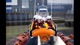 Taken from the bow of an orange lifeboat, three women crew members sit in the lifeboat, wearing white helmets, red lifejackets and yellow and black drysuits.
