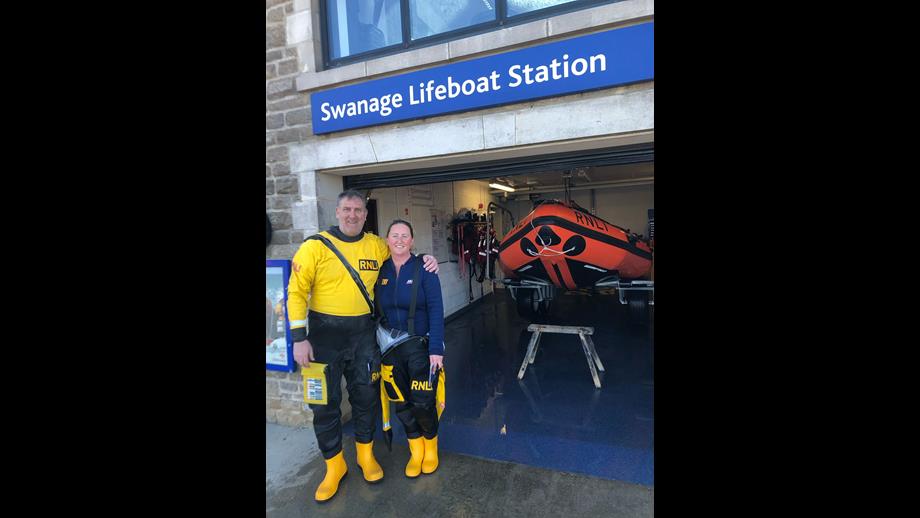 Man and woman wearing yellow and black drysuit gear stand in entryway to lifeboat station. Orange lifeboat is in background.
